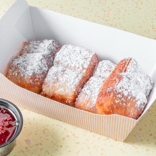 Powdered sugar beignets in a striped box with a side of red jam on a speckled yellow surface.