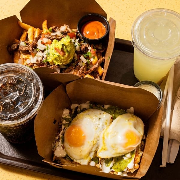 Two takeout containers with loaded fries, chilaquiles with fried eggs, and two drinks on a tray.