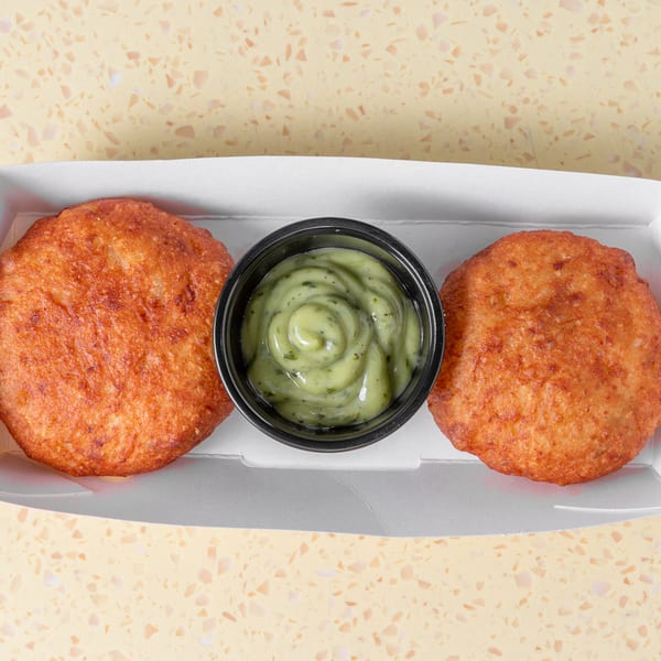 Two golden-brown fried patties and a cup of green creamy sauce in a white takeaway box.