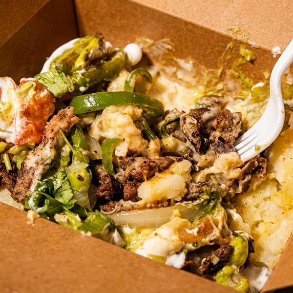 Loaded baked potato with meat, cheese, peppers, and sauce in a brown takeout container with a fork.