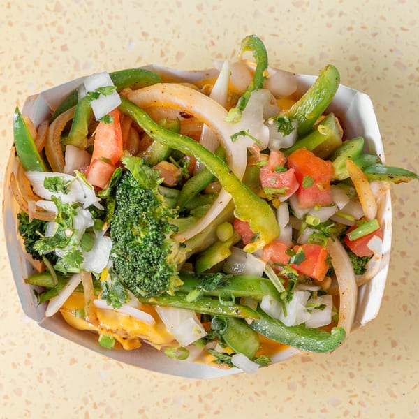 Chopped broccoli, green peppers, onions, and tomatoes in a white paper container with cheese sauce.