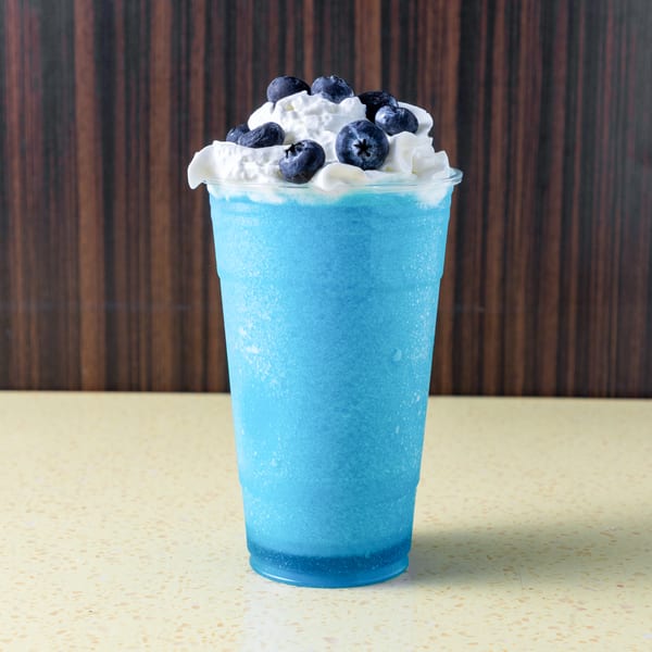 Blue frozen drink with whipped cream and blueberries in a clear cup on a speckled counter.