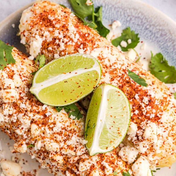Two elote (Mexican street corn) ears topped with cheese, chili powder, lime wedges, and cilantro.
