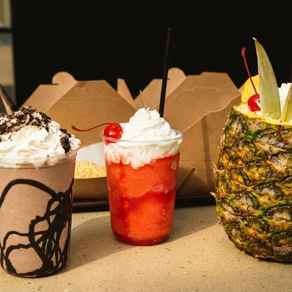 Three vibrant drinks: a chocolate shake, a red slushie, and a pineapple drink, on a counter.
